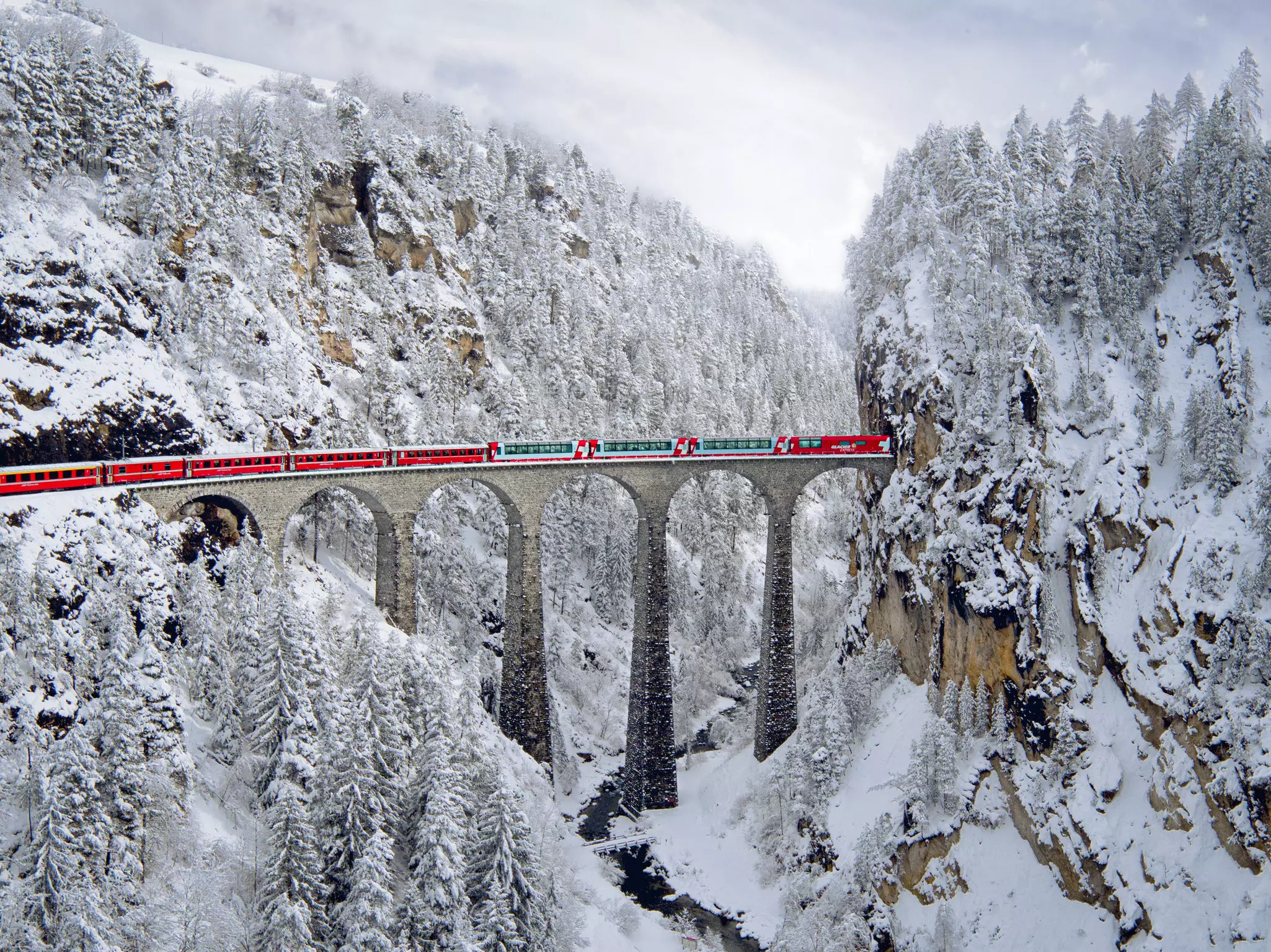 A red train crosses a six-arched bridge across a snowy mountain valley.