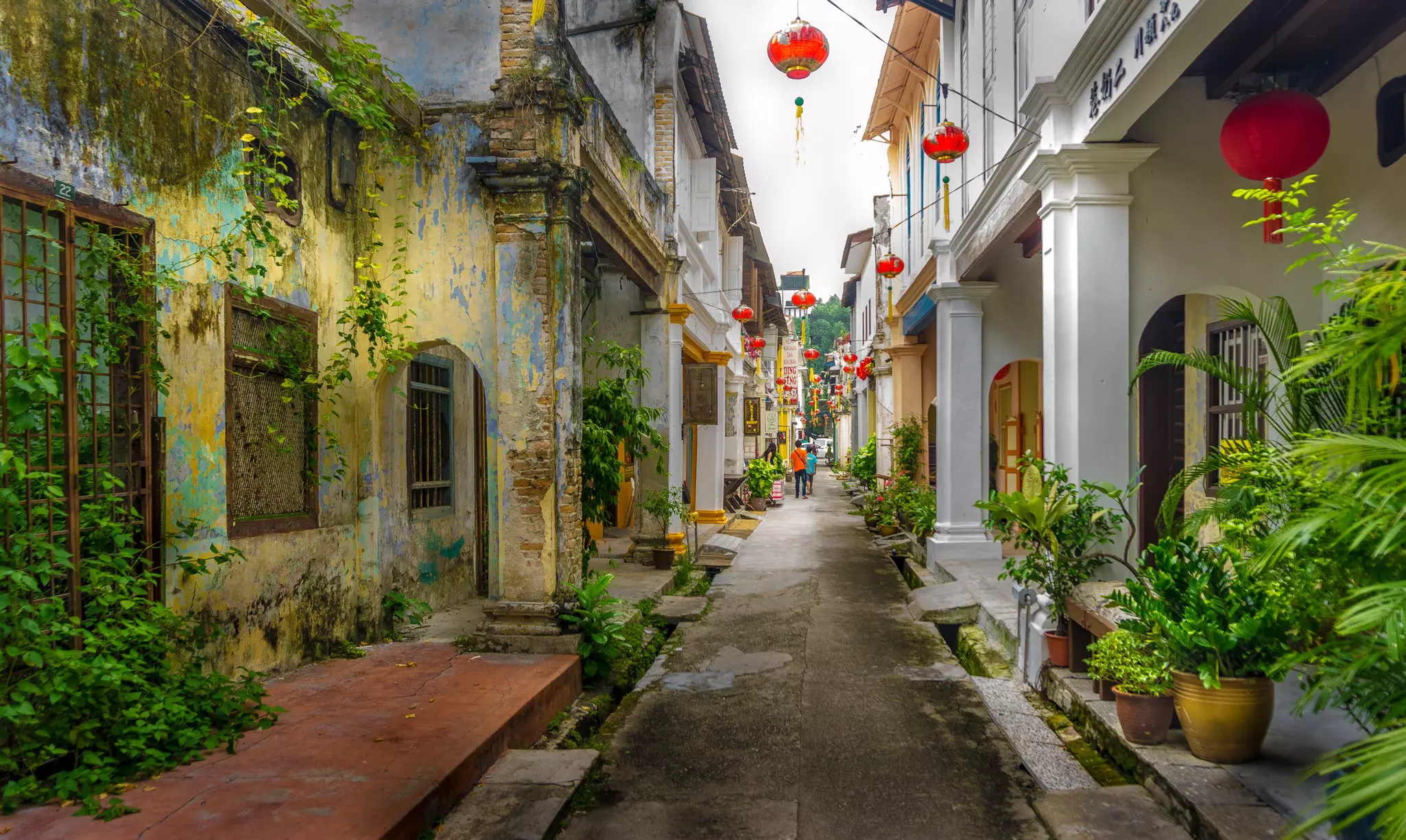 A narrow old street with red Chinese-style paper lanterns strung between the buildings. Two figures are walking away from the camera