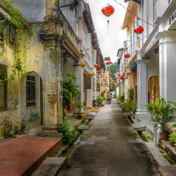 A narrow old street with red Chinese-style paper lanterns strung between the buildings. Two figures are walking away from the camera