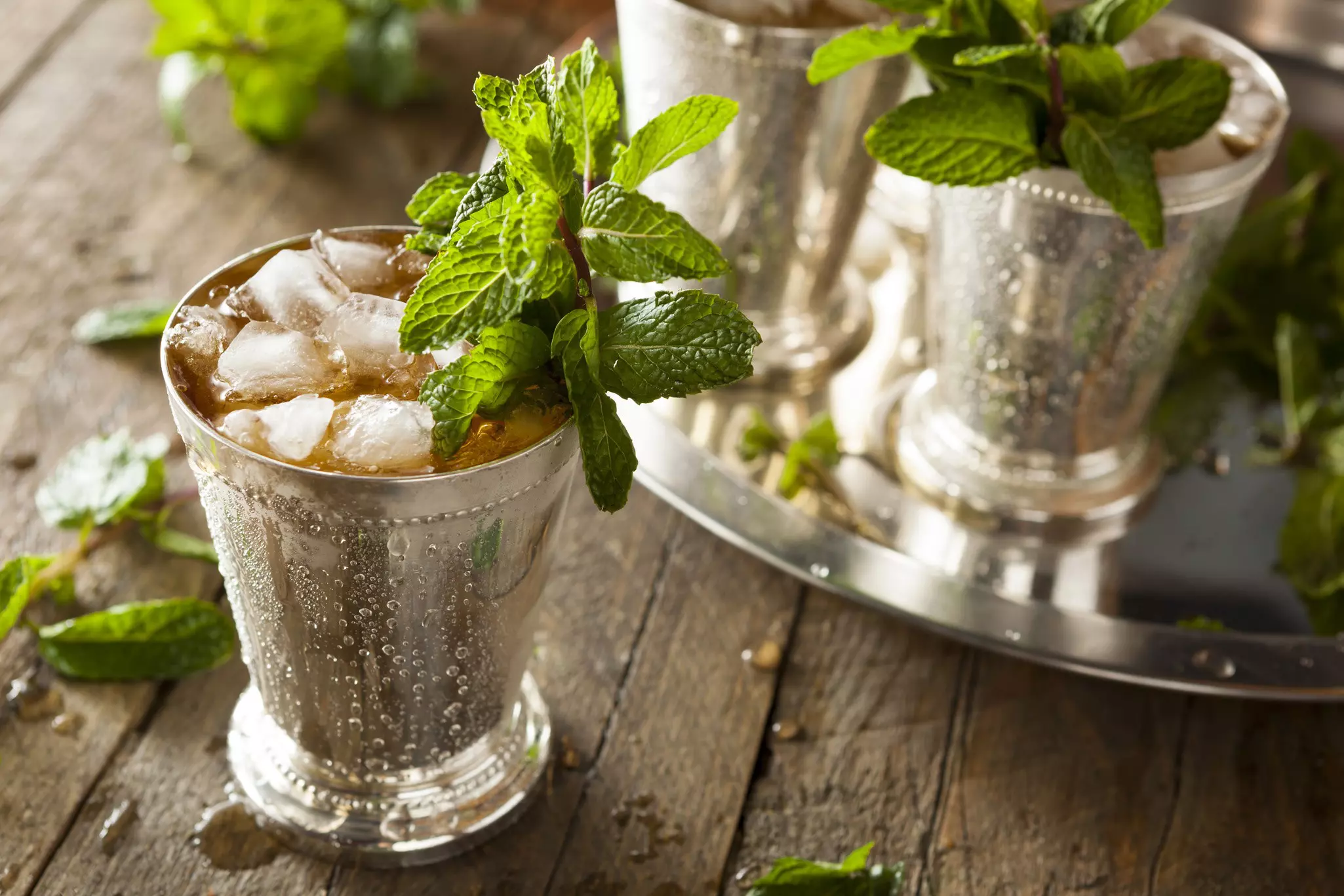 Close-up of silver glass containing brown liquor, ice, and mint sprigs on a table with similar glasses in the background.