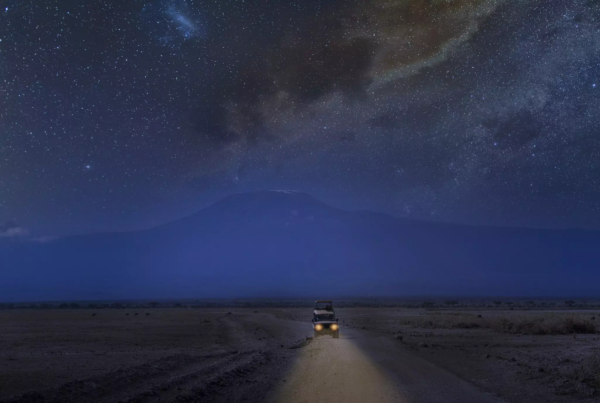 A 4WD truck follows a gravel track in a national park at night with the silhouette of a large pointed volcano looming in the distance