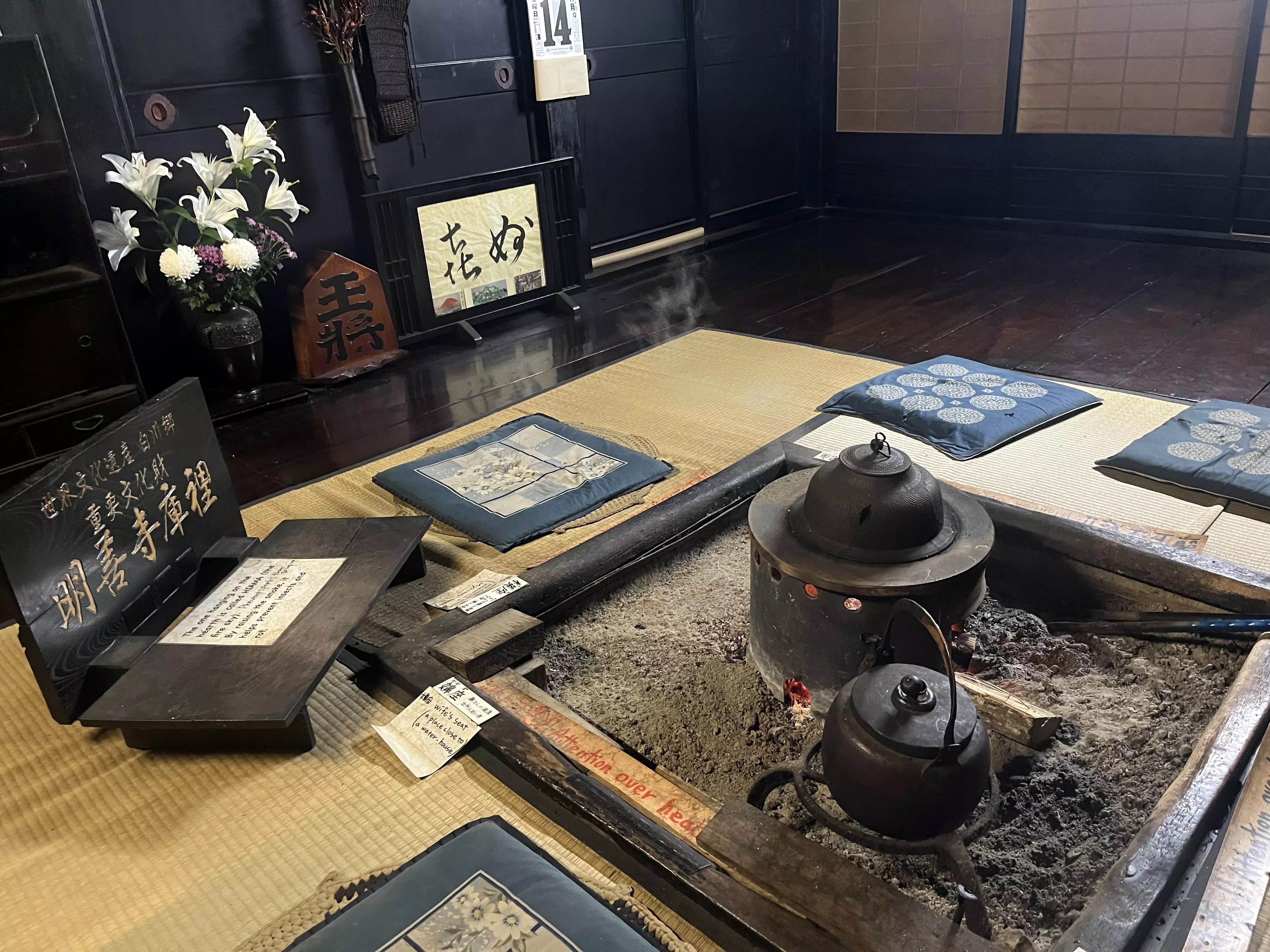 A teapot inset into a table with ashes, surrounded by pillows and low seating with flowers and  a black wall in the distance.