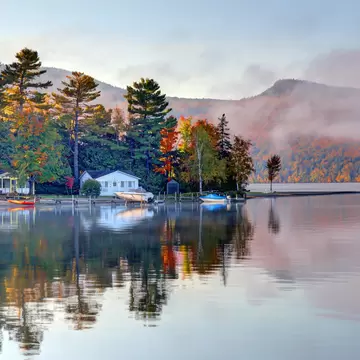 Fall foliage surrounding a lake. Small, wooden houses sit on the shore with small boats docked nearby: Lake Willoughby, Vermont