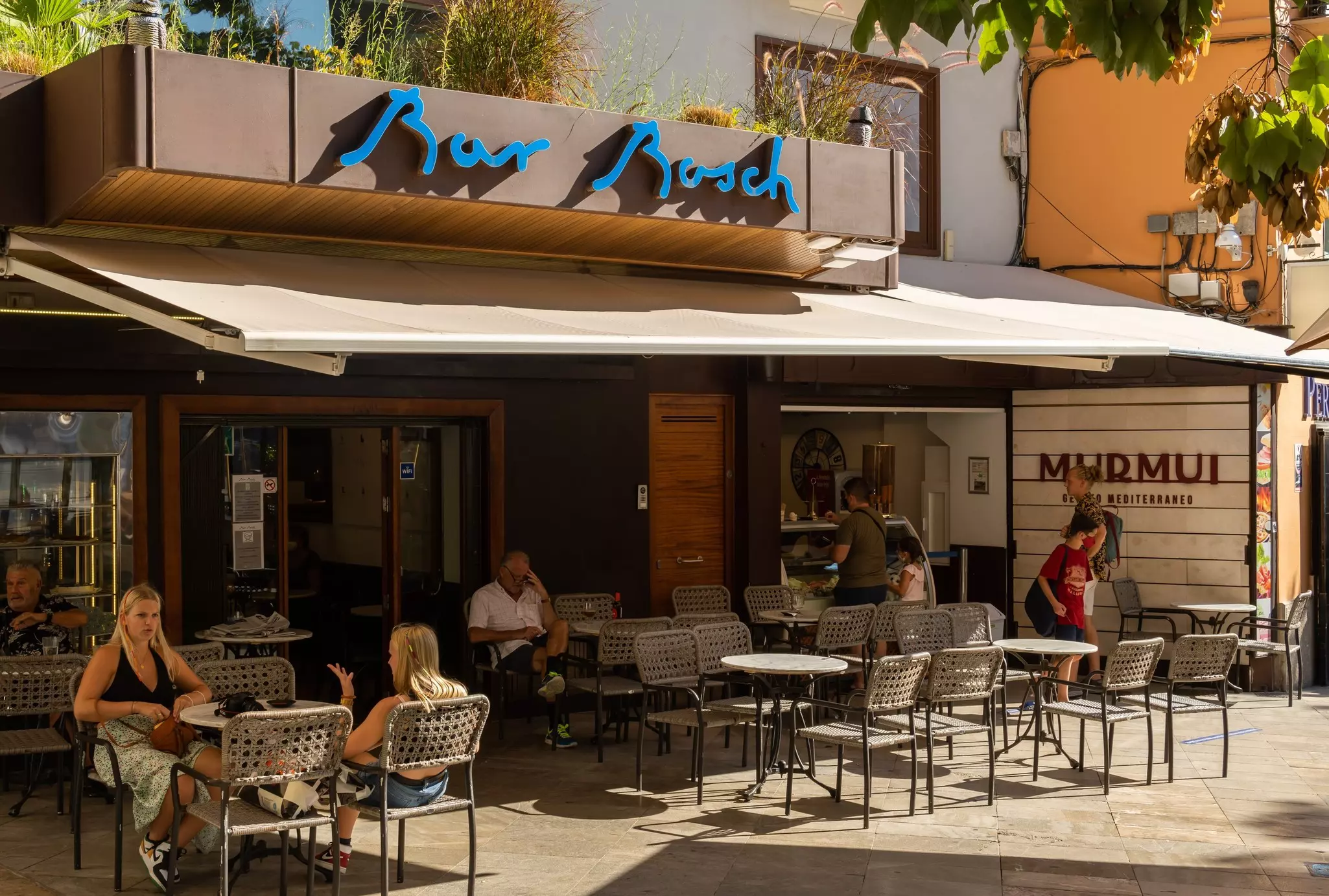 People sit outside at tables under an awning at a bar in Spain.