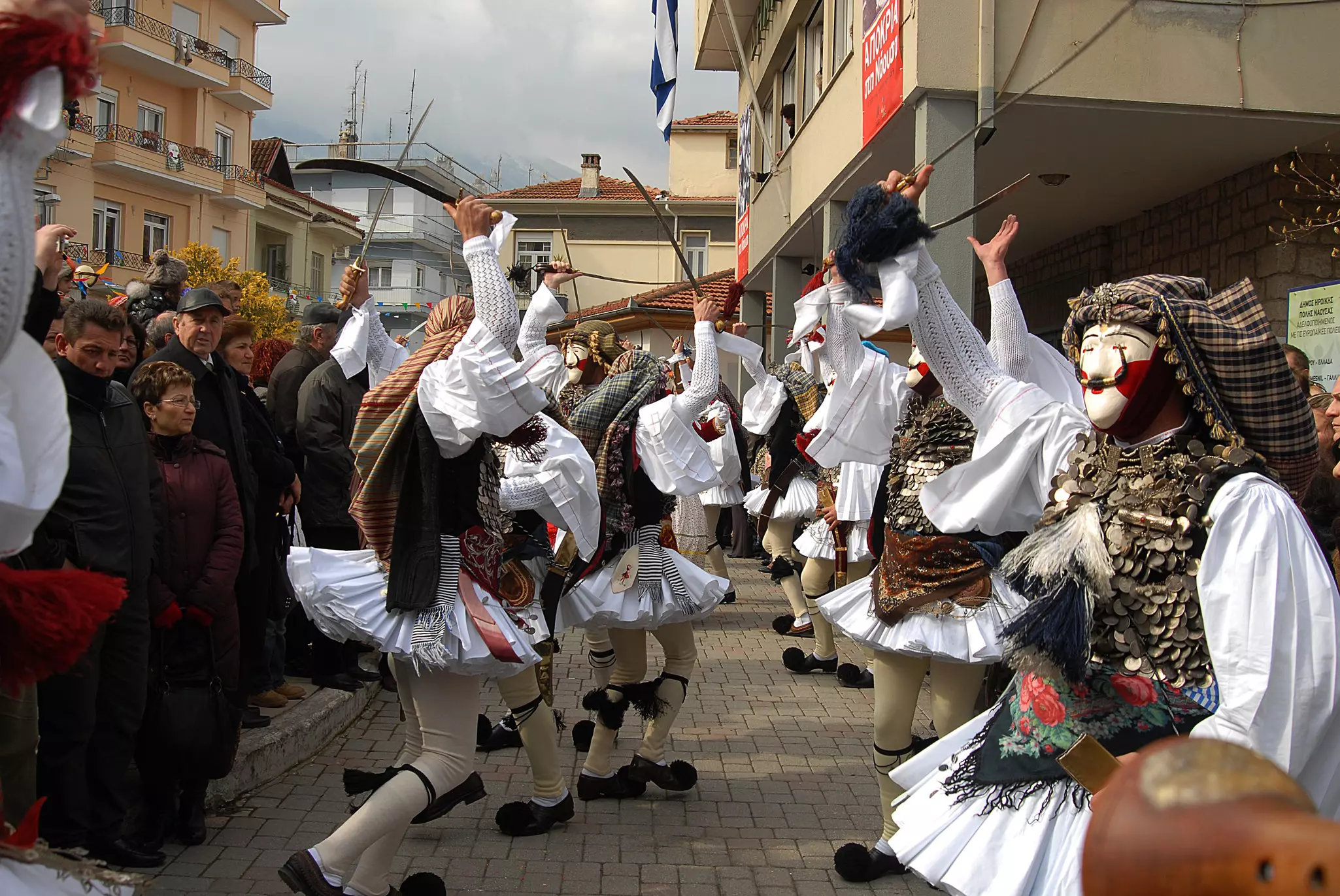People in traditional folk costumes and masks dance in the street of a village during a daytime celebration.