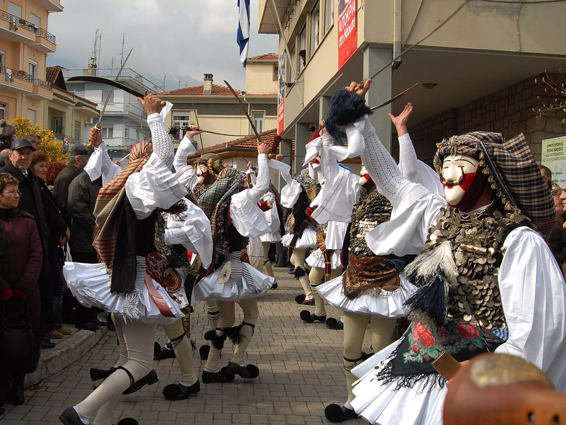 People in folk clothing and masks dance in the street of a village during a daytime celebration.
