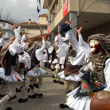 People in folk clothing and masks dance in the street of a village during a daytime celebration.