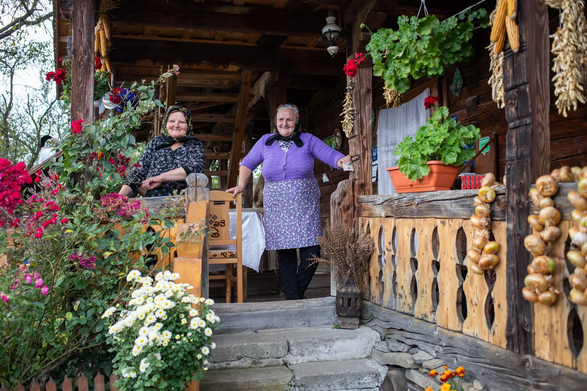 Two older women wearing headscarves stand on the porch of a traditional wooden house decorated with flower boxes. Bulbs of garlic are draped over a wooden railing.
