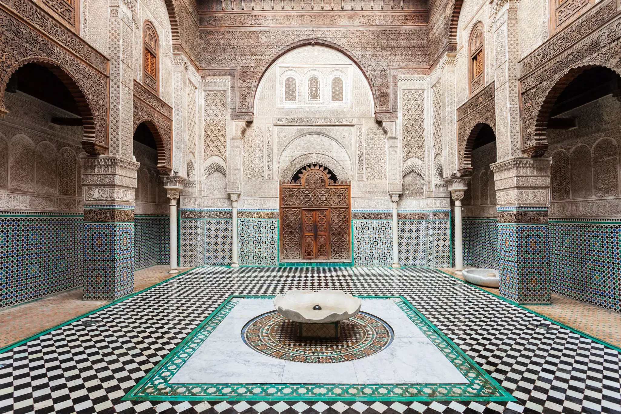 A courtyard in a building with detailed geometric tilework covering the floor, walls and surrounding a carved wooden doorway.