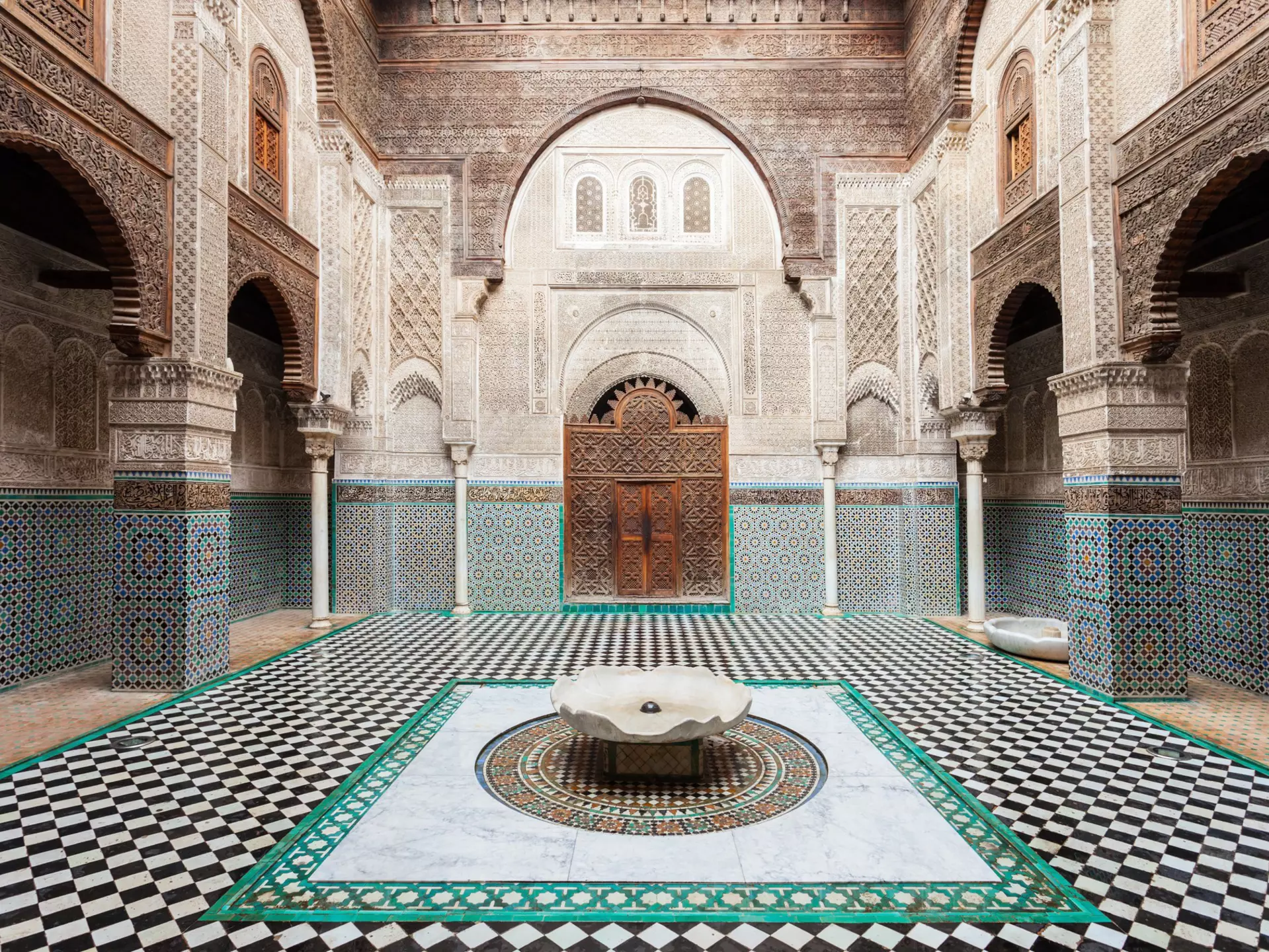 A courtyard in a building with detailed geometric tilework covering the floor, walls and surrounding a carved wooden doorway.