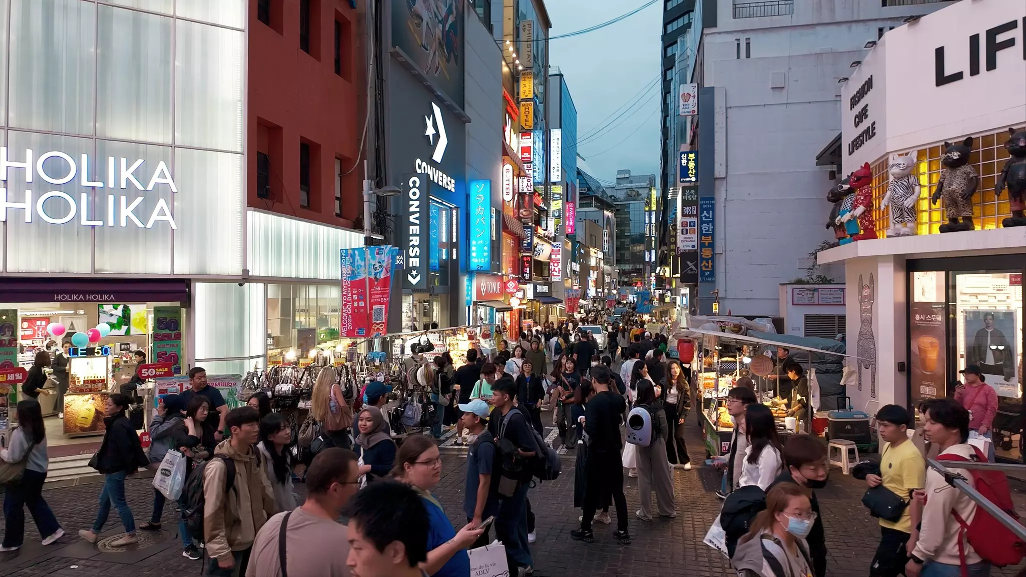 Crowds of people walk in a pedestrianized street at dusk. The lights of stores and signs illuminate the scene.