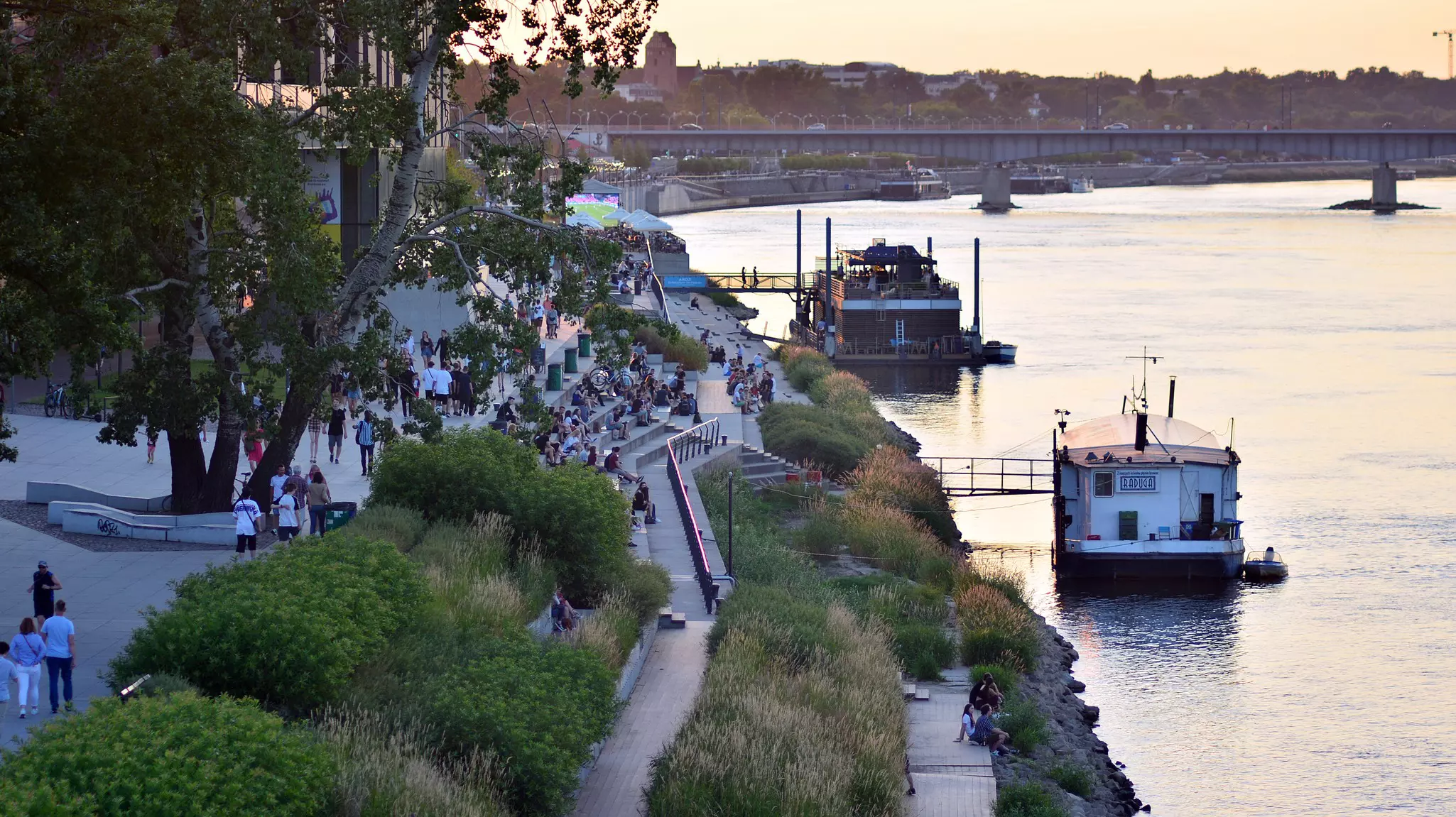 People walking and sitting along a landscaped walkway by the river in Warsaw, Poland, at dusk; there are ferry boats docked by the shore.