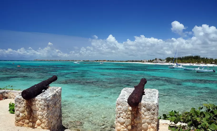 Two old cannons on coral-stone mounts overlooking a turquoise Caribbean bay dotted with boats