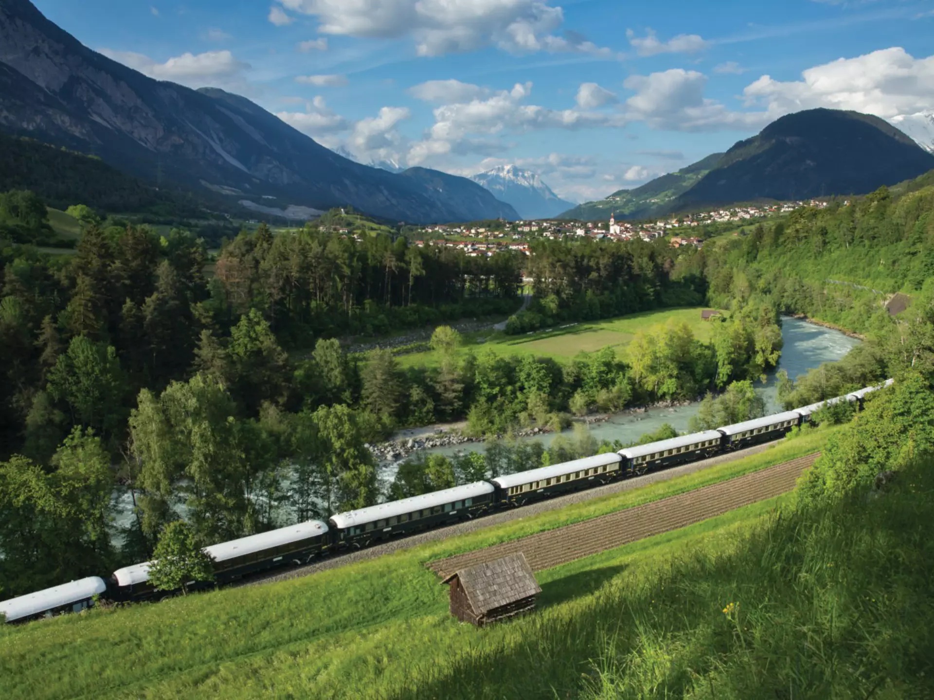 A train travels through a green valley. A town and snowcapped mountains are in the distance.