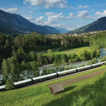 A train travels through a green valley. A town and snowcapped mountains are in the distance.
