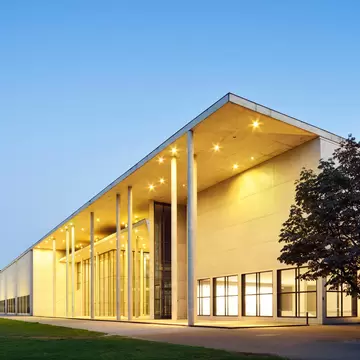 Dusk shot of a vast white modern building with large square windows and thin columns supporting the roof. There are a couple of trees to the left of the building.