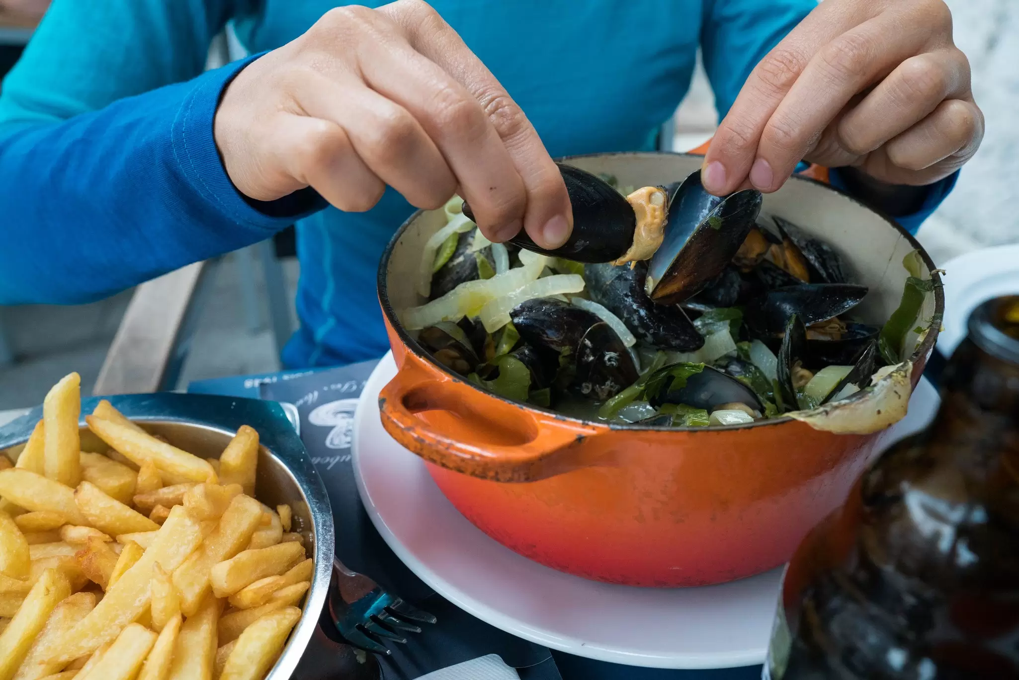 Hands holding two mussels shells over an orange pot, with a plate of fries to the side.
