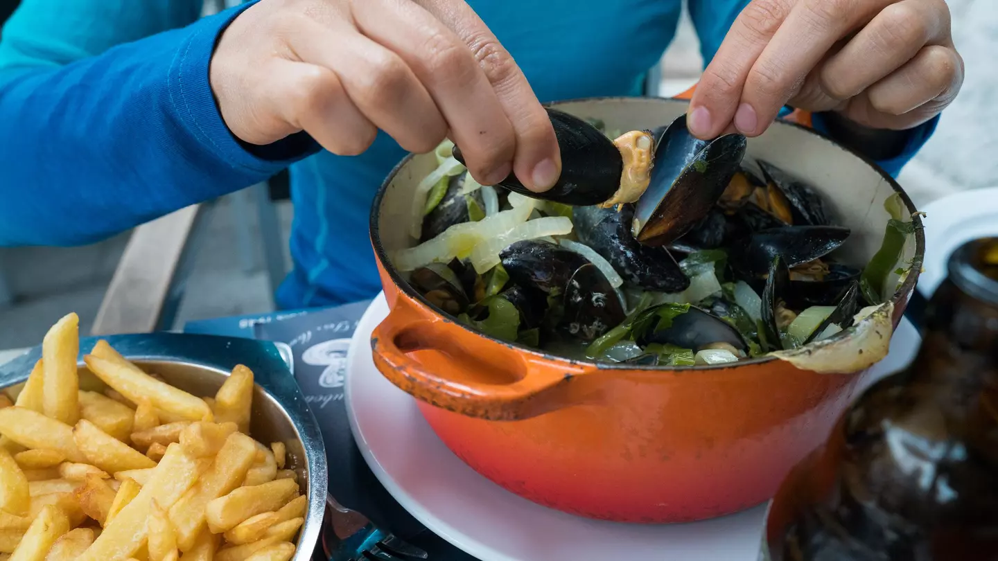 Hands holding two mussels shells over an orange pot, with a plate of fries to the side.