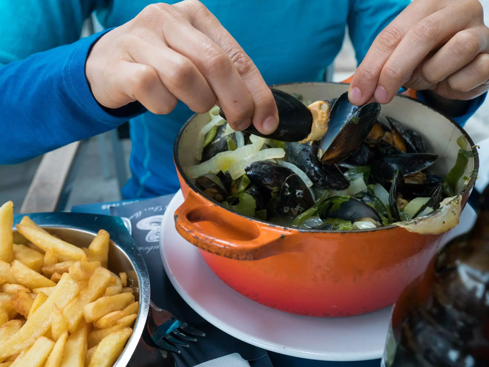 Hands holding two mussels shells over an orange pot, with a plate of fries to the side.