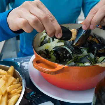 Hands holding two mussels shells over an orange pot, with a plate of fries to the side.