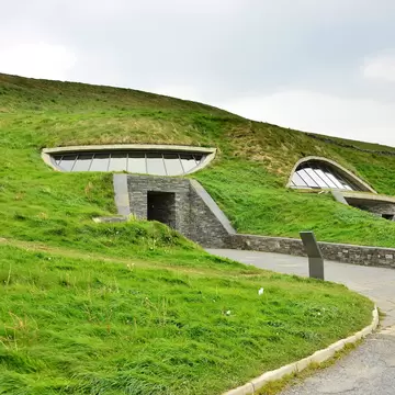 A grass-covered hillside with curved glass windows and a slate stone entrance set into it, a paved path curving past.
