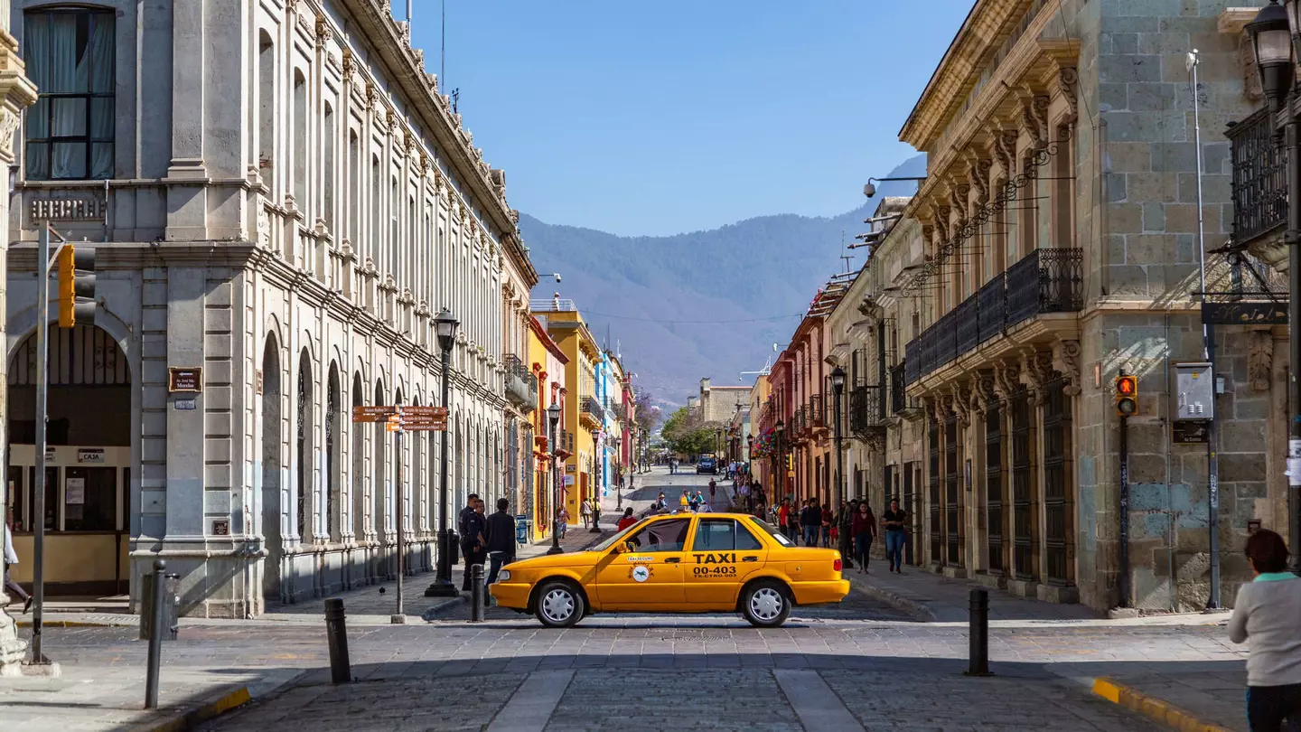 A yellow taxi drives across a cobbled street lined with historic buildings.
