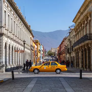 A yellow taxi drives across a cobbled street lined with historic buildings.