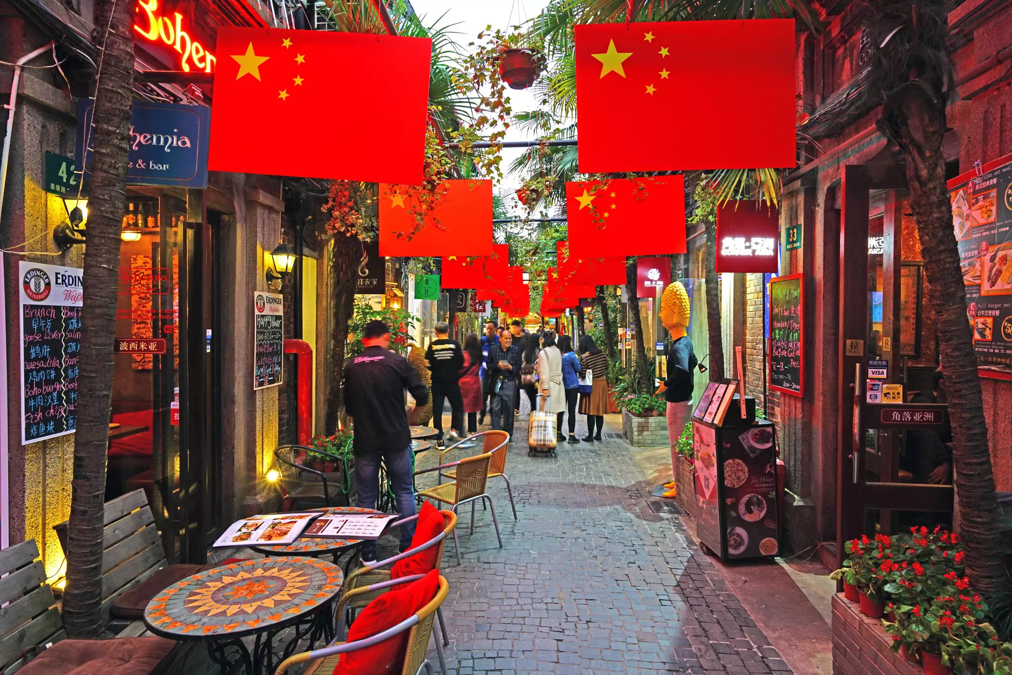 A narrow alley lined with cafes and restaurants and decorated with many red Chinese flags.