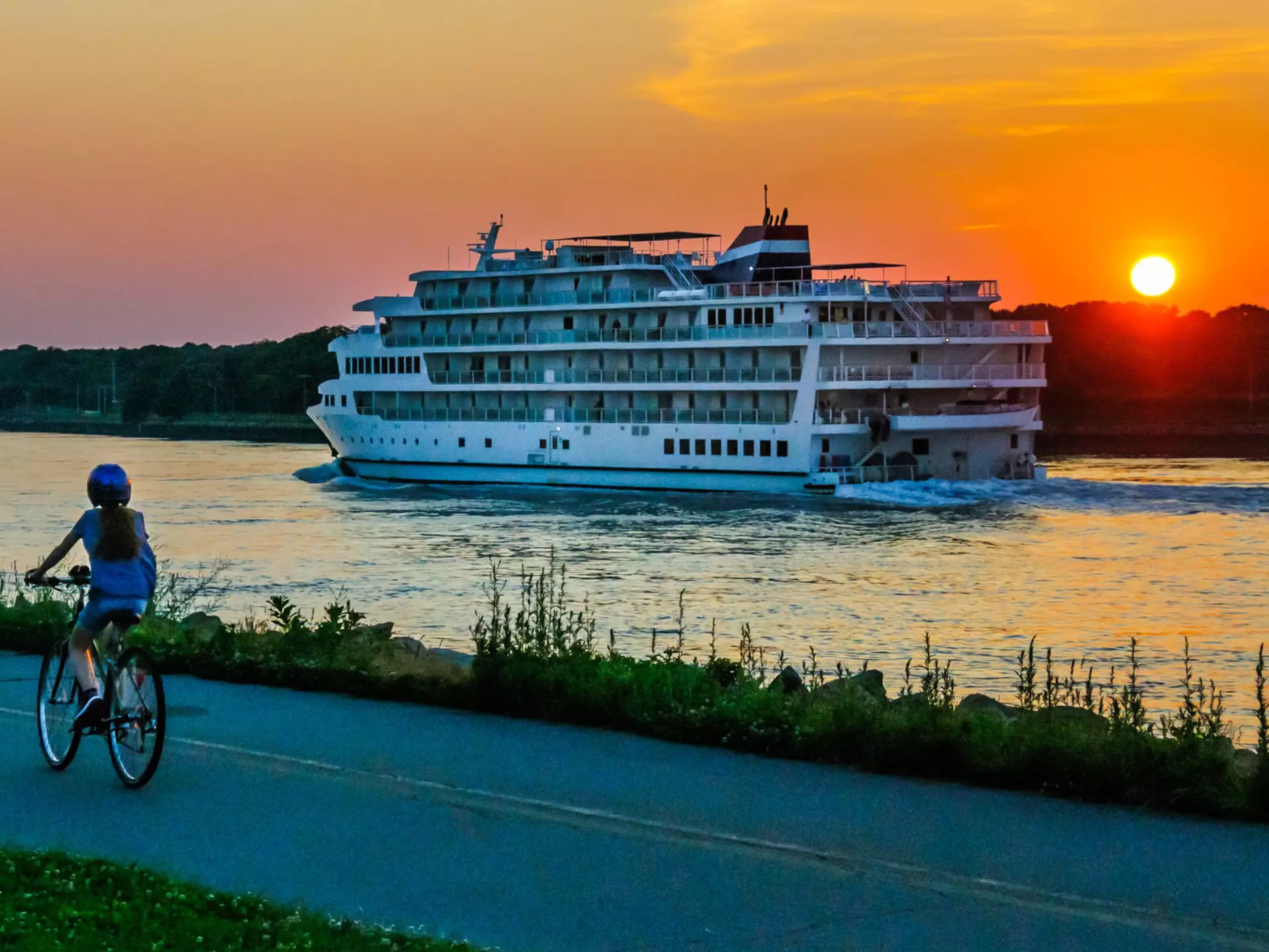 A young woman on a bicycle watches a cruise ship on the Cape Cod Canal