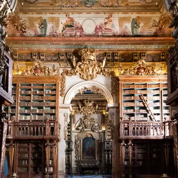 Baroque library hall with gilded wooden bookcases, frescoed ceiling and a red carpet leading to a royal portrait, Coimbra.
