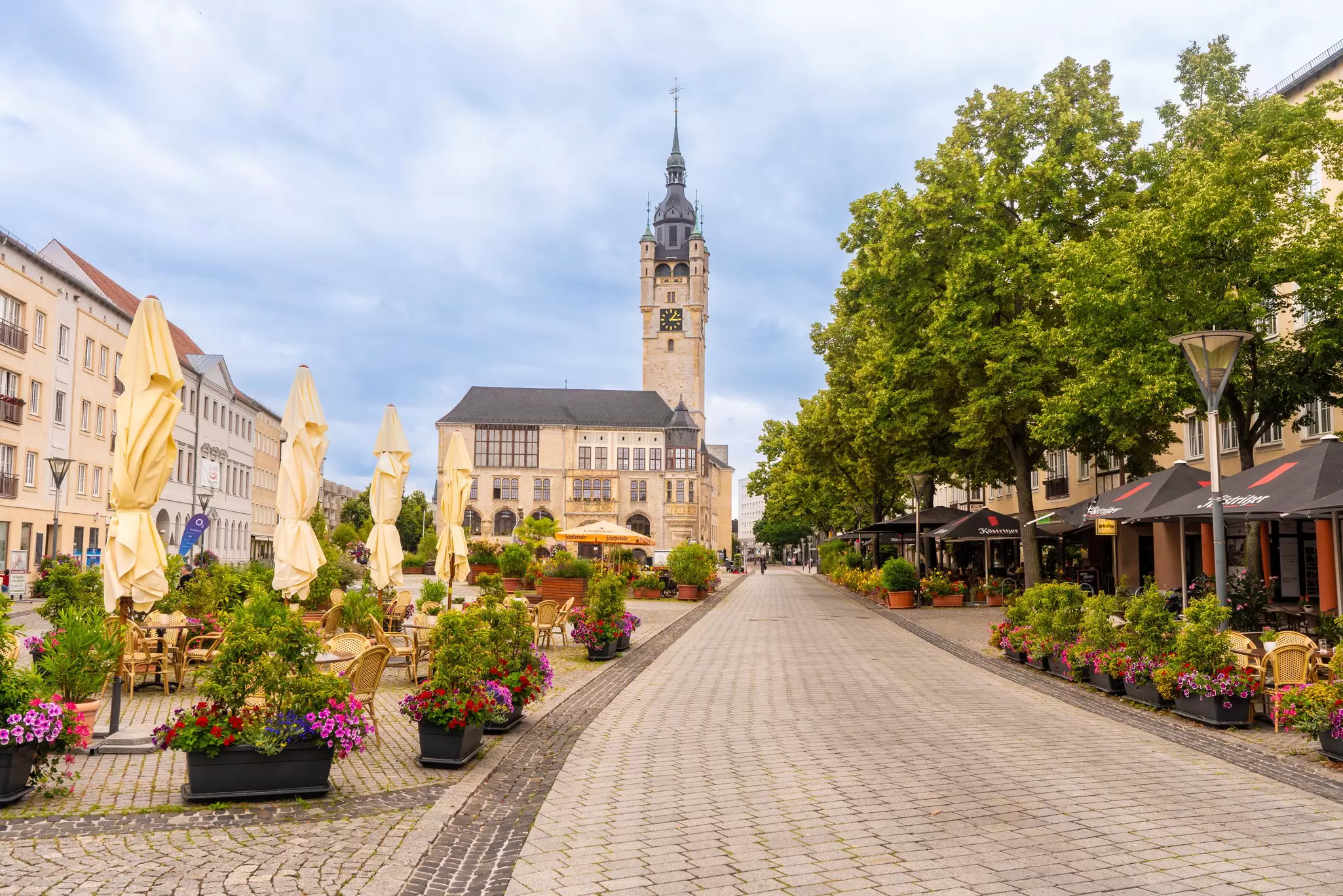 A town square lined with cafes, colorful flowers, and a prominent clock tower of the city hall under a cloudy sky