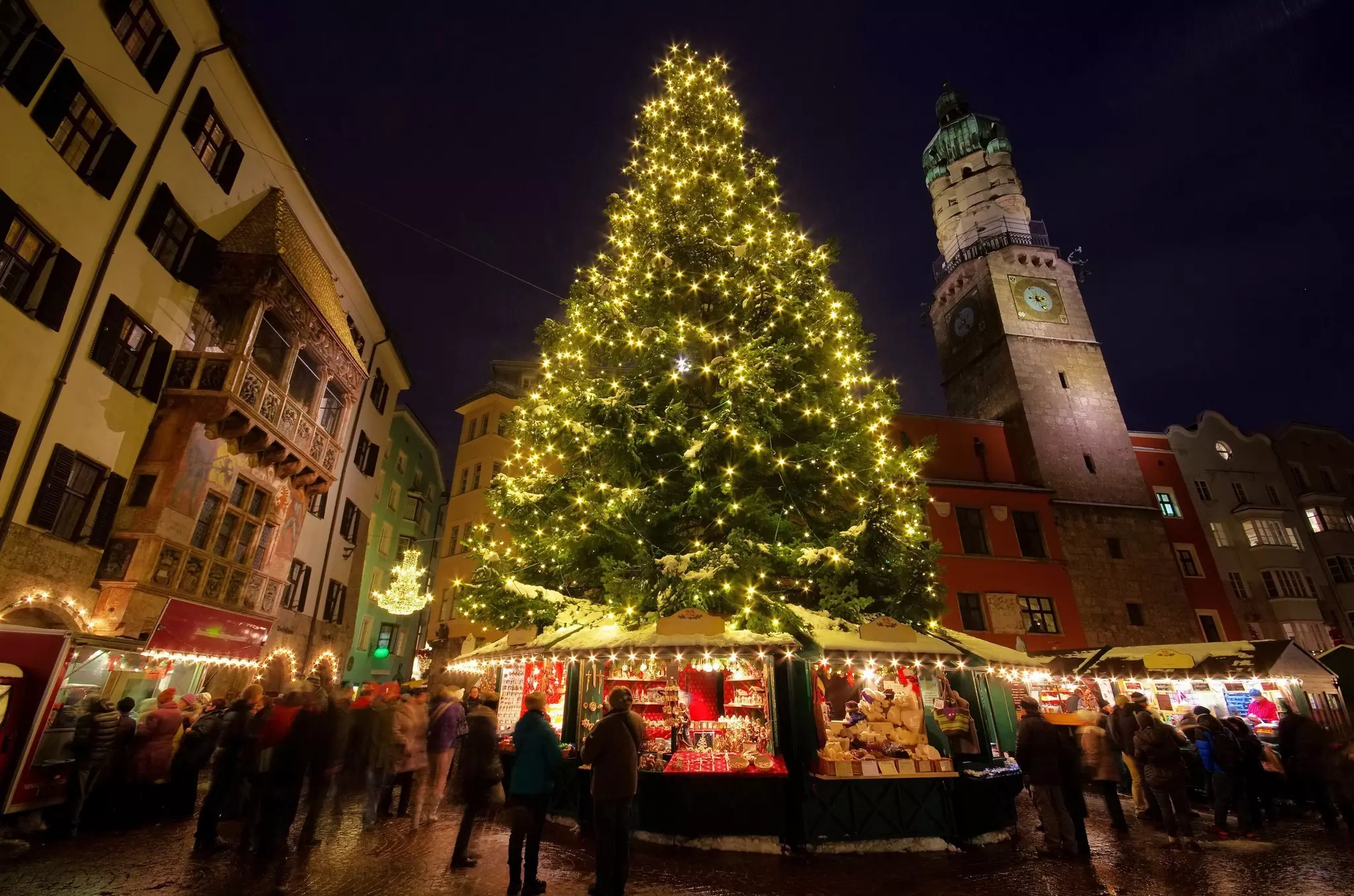 A large Christmas tree stands among market stalls in a city center lined with medieval buildings.
