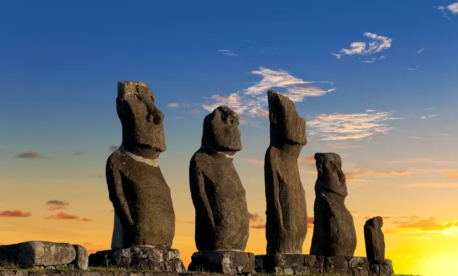 Five moai statues on a stone ahu platform on Rapa Nui (Easter Island), silhouetted against an orange sunset and blue sky