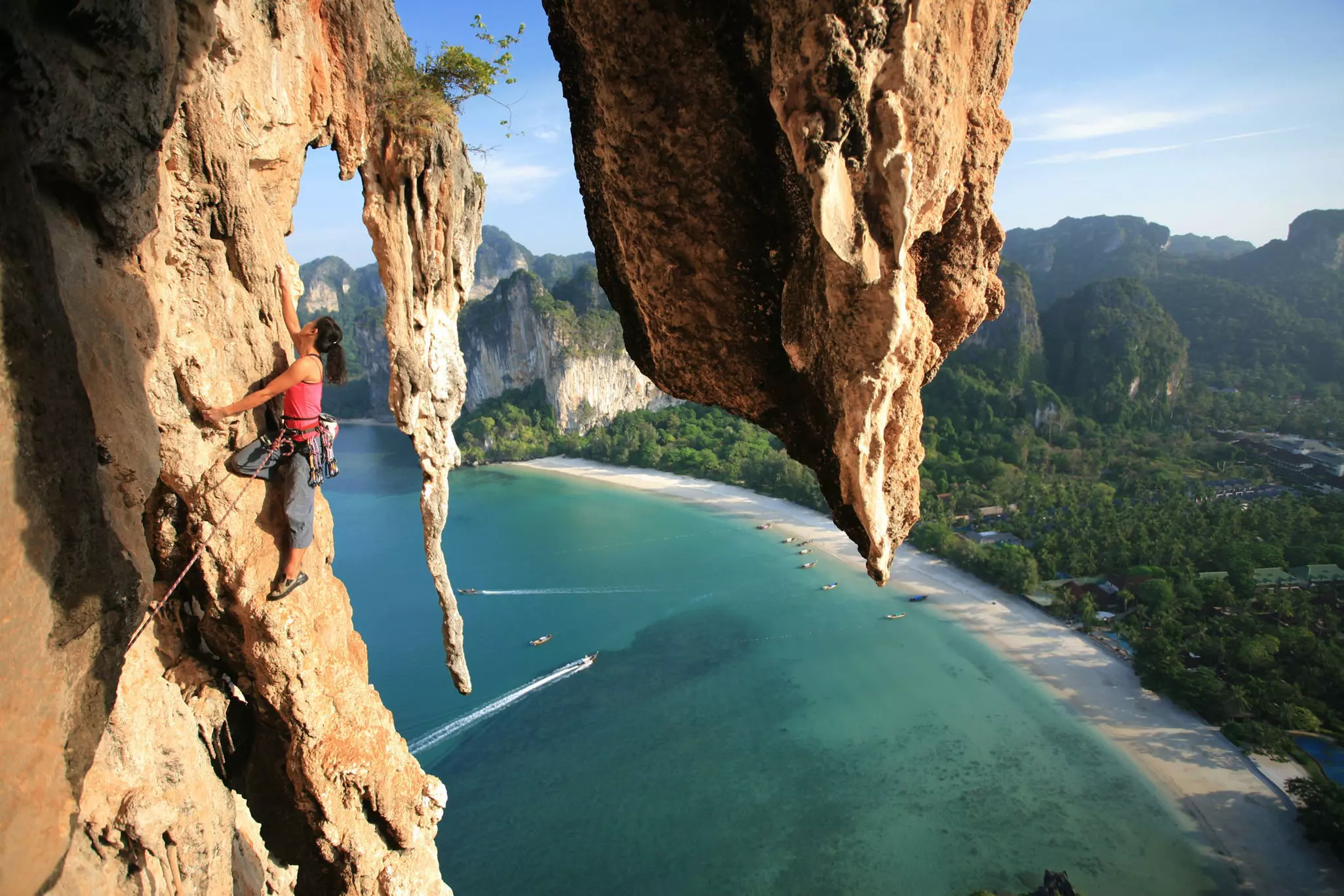 A rock climber clings to a rock face high above a tropical beach.