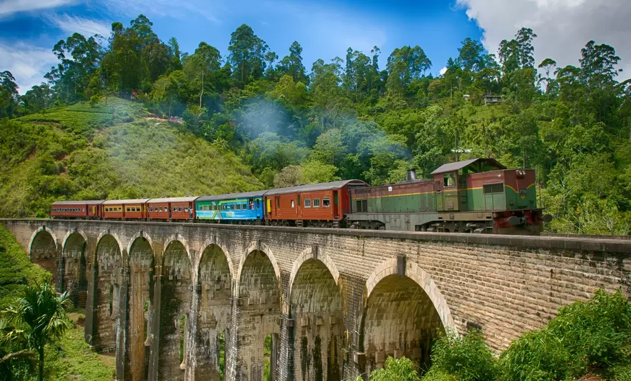 Train with a green diesel engine and red and blue carriages crossing the stone Nine Arch Bridge near Ella, Sri Lanka