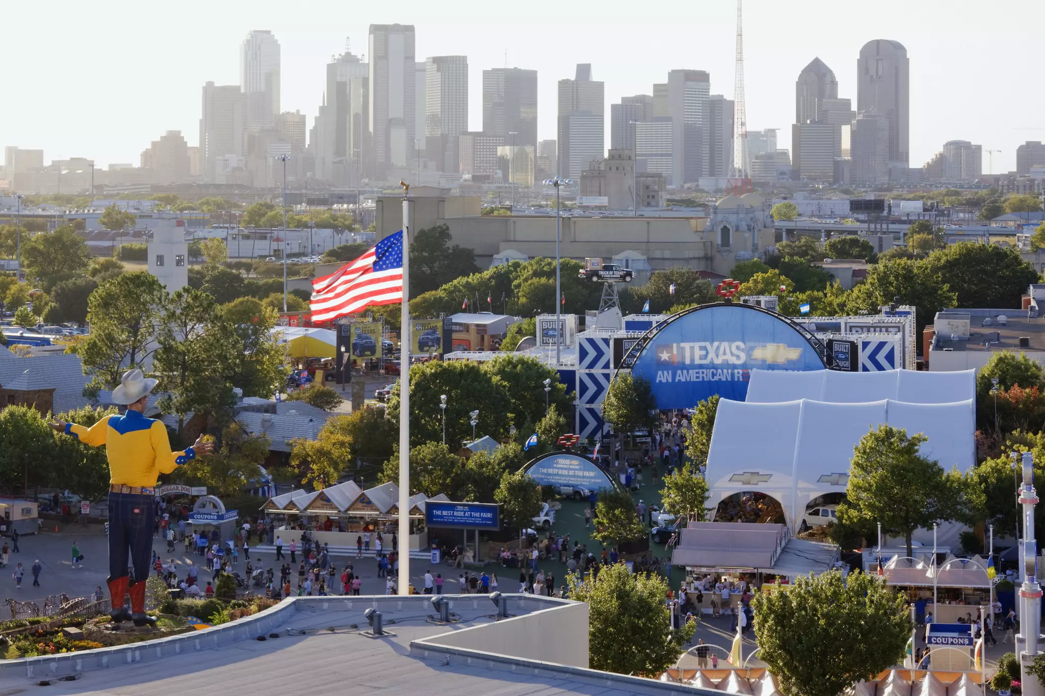 An American flag flies near a giant cowboy statue in front of a massive fair.