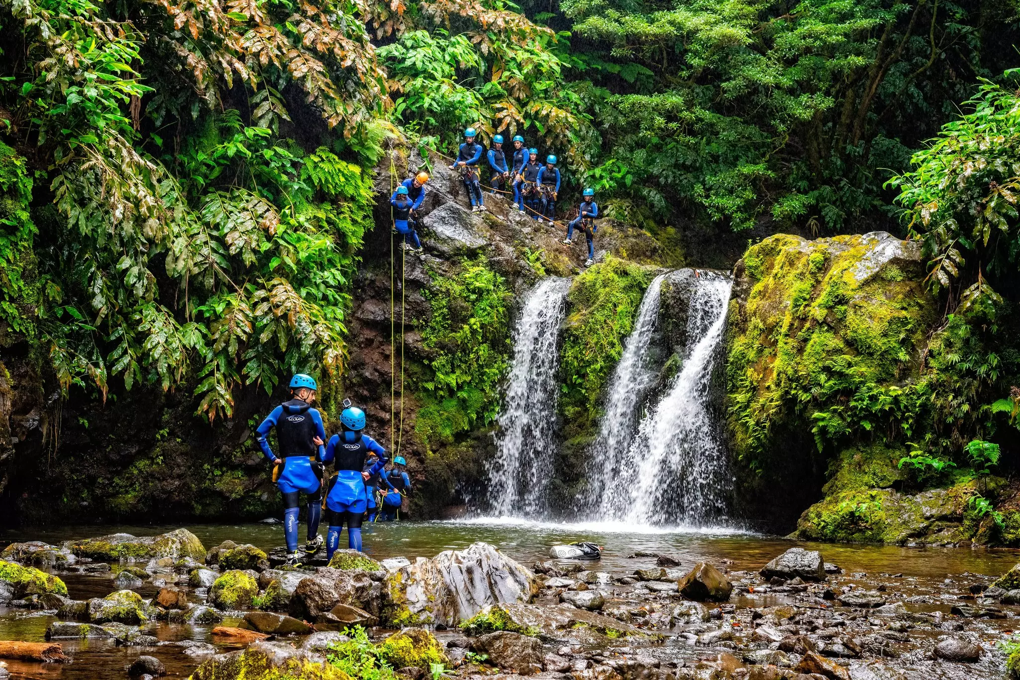 People standing on slick rocks on either side of a pool of water by a waterfall; they are wearing canyoning gear, and there are yellow ropes attached to the top of the rocks that extend to the water.