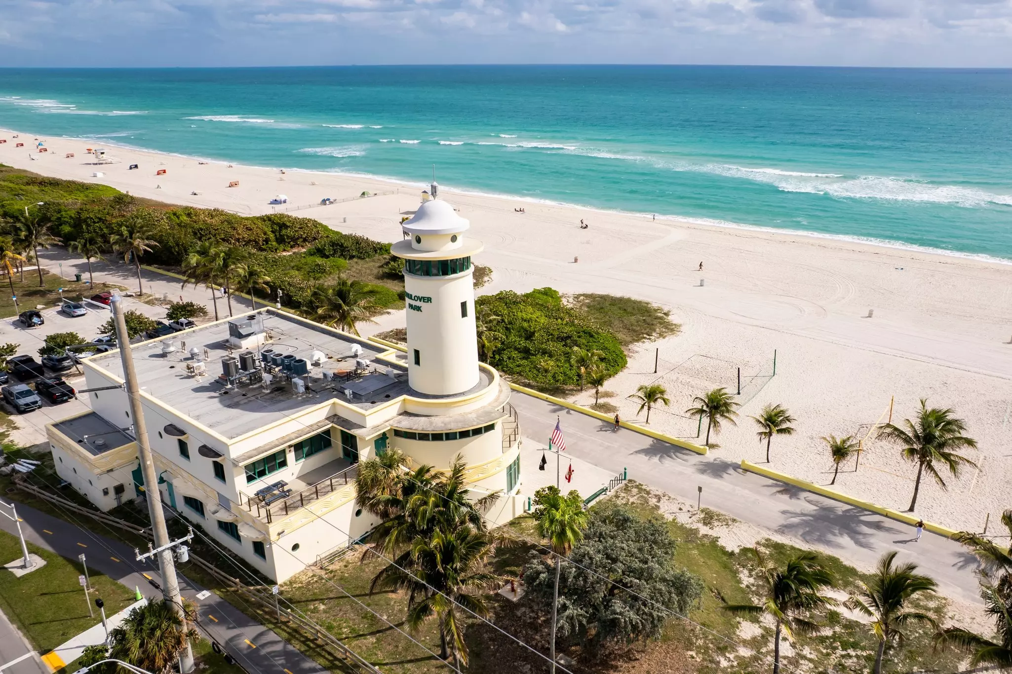 An aerial view of a lighthouse in front of a wide beach lined by palms.