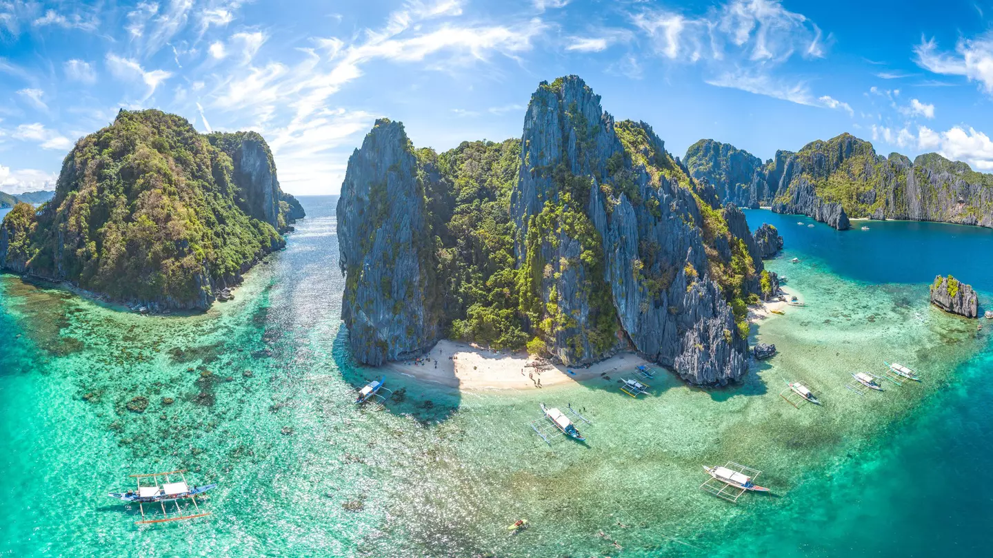 An aerial shot of El Nido, Palawan Province, Philippines. The small islands have very high rocky cliffs and lots of dense greenery. There are some white sandy beaches and a few boats moored near the islands.