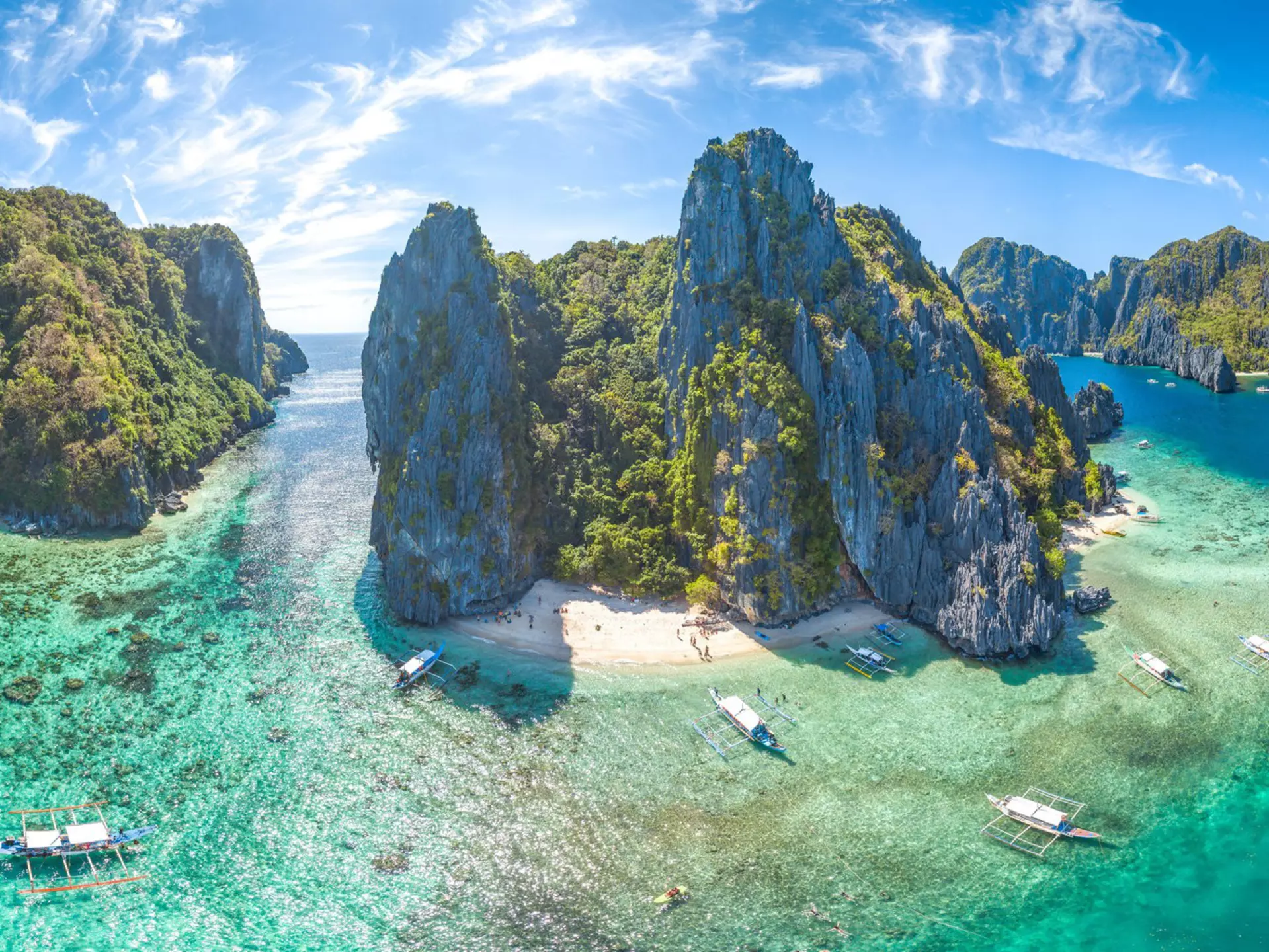 An aerial shot of El Nido, Palawan Province, Philippines. The small islands have very high rocky cliffs and lots of dense greenery. There are some white sandy beaches and a few boats moored near the islands.
