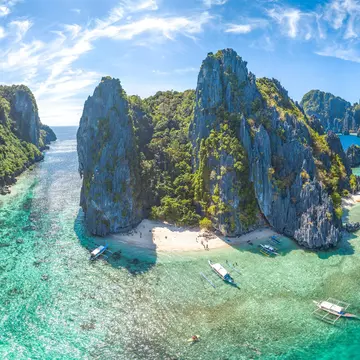 An aerial shot of El Nido, Palawan Province, Philippines. The small islands have very high rocky cliffs and lots of dense greenery. There are some white sandy beaches and a few boats moored near the islands.