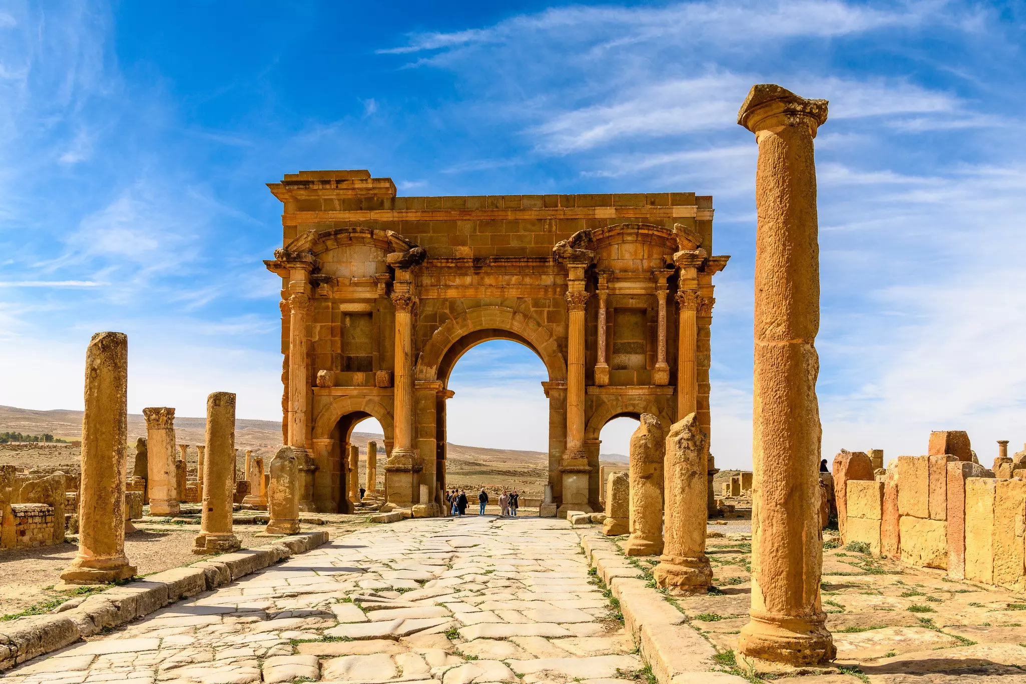 An ancient Roman archway with people standing below it at the end of a stone pathway lined with columns.