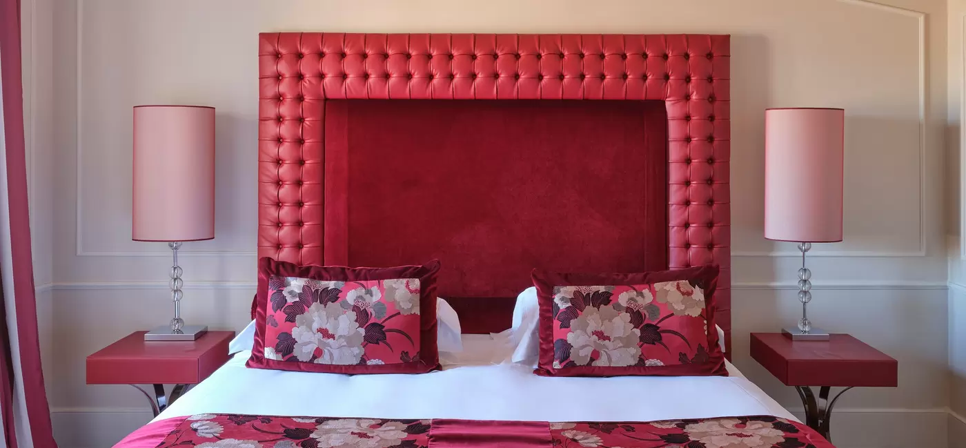 A hotel room with a bed with a red headboard, red floral bedding, red side tables with red lamps on either side