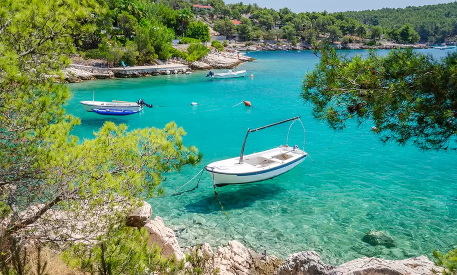 Small boats moored in a clear turquoise cove fringed by pine trees and white limestone rocks on Brač Island, Croatia