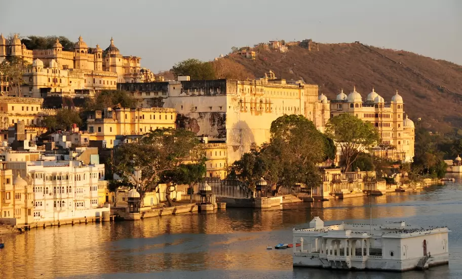 Udaipur's City Palace in golden evening light above Lake Pichola, a small white pavilion in the water and hills behind