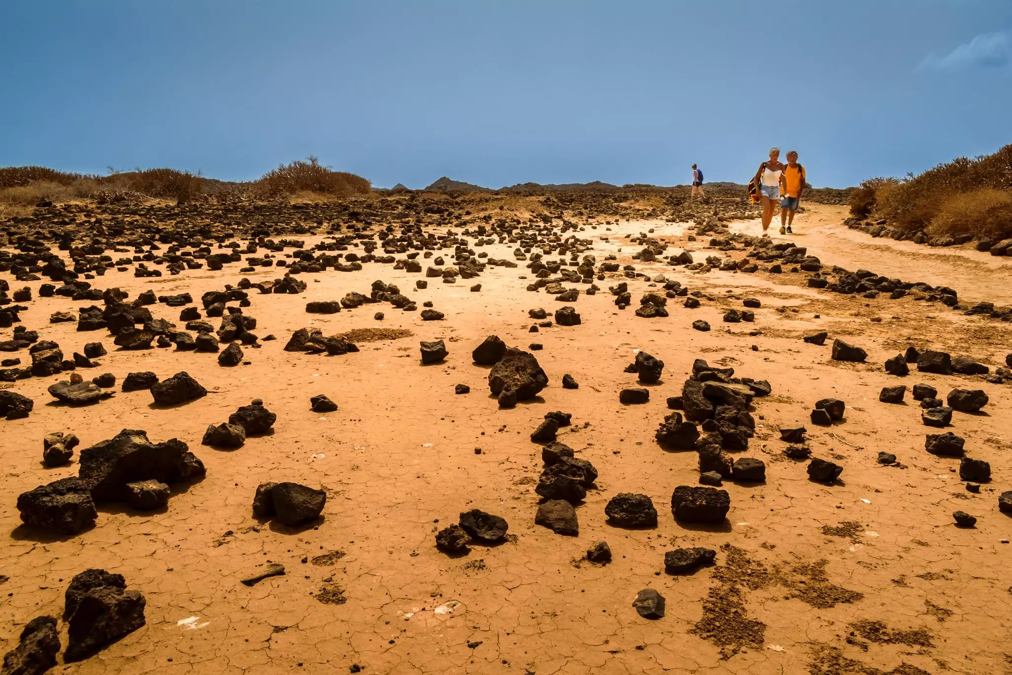 Rocks amid sand with two people walking nearby and dunes in the background