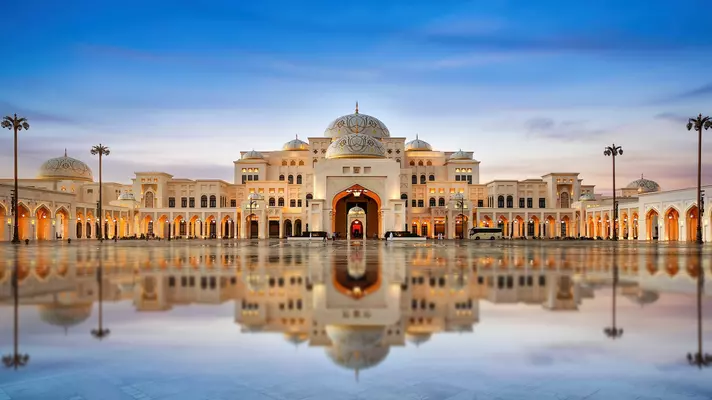 A view of an ornate palace with a symmetrical facade reflected in a shallow pool in front. Domes and archways cover the building.