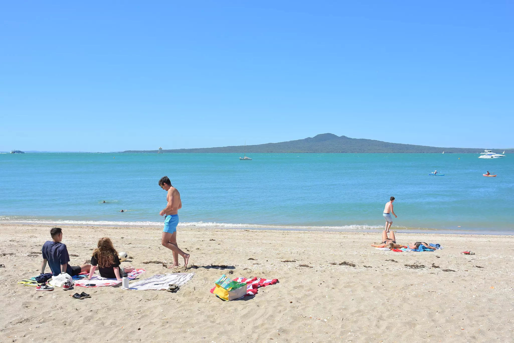 People sunbathing on Mission Bay beach with Rangitoto Island in the background.