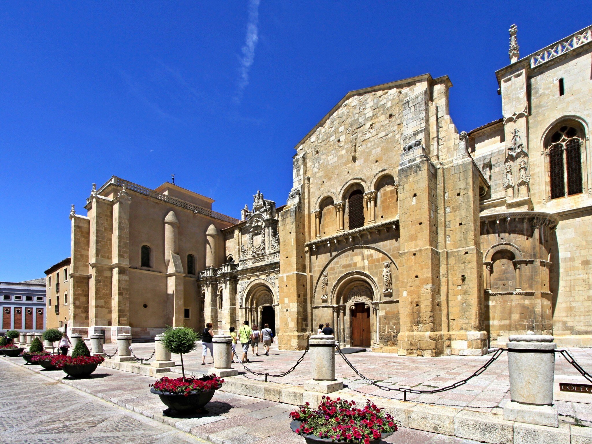 A honey-stone church front with a carved arched doorway and a later Gothic wing, across a paved square set with flower planters.
