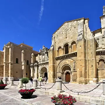 A honey-stone church front with a carved arched doorway and a later Gothic wing, across a paved square set with flower planters.